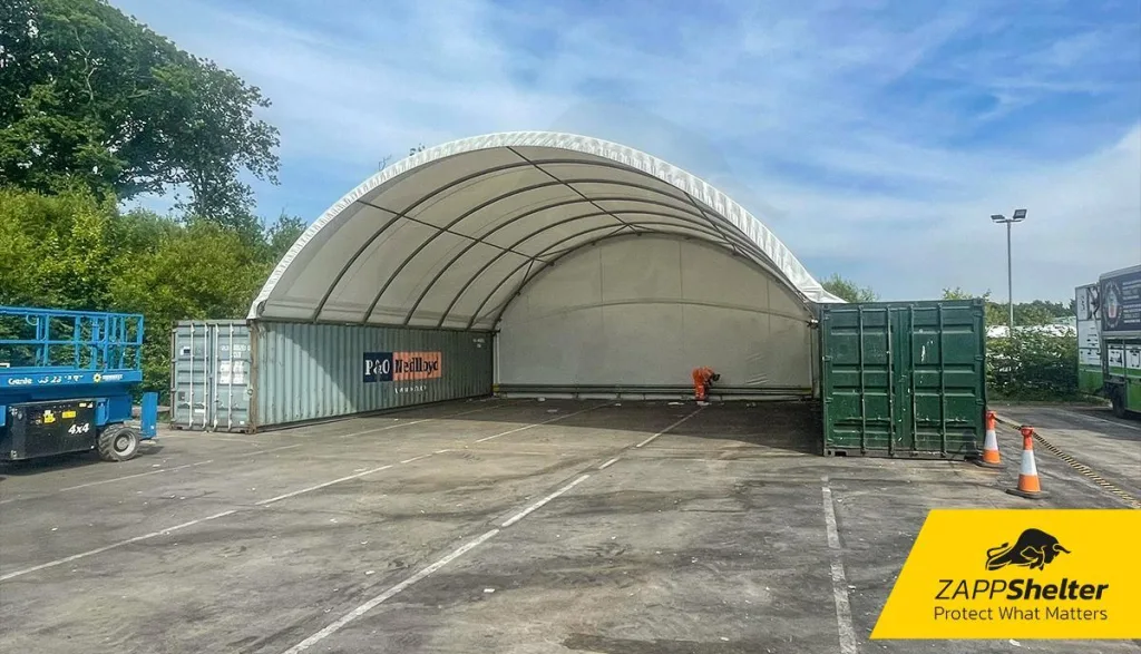 A white Zappshelter fabric canopy with a rear end wall, mounted on green shipping containers in a paved yard.